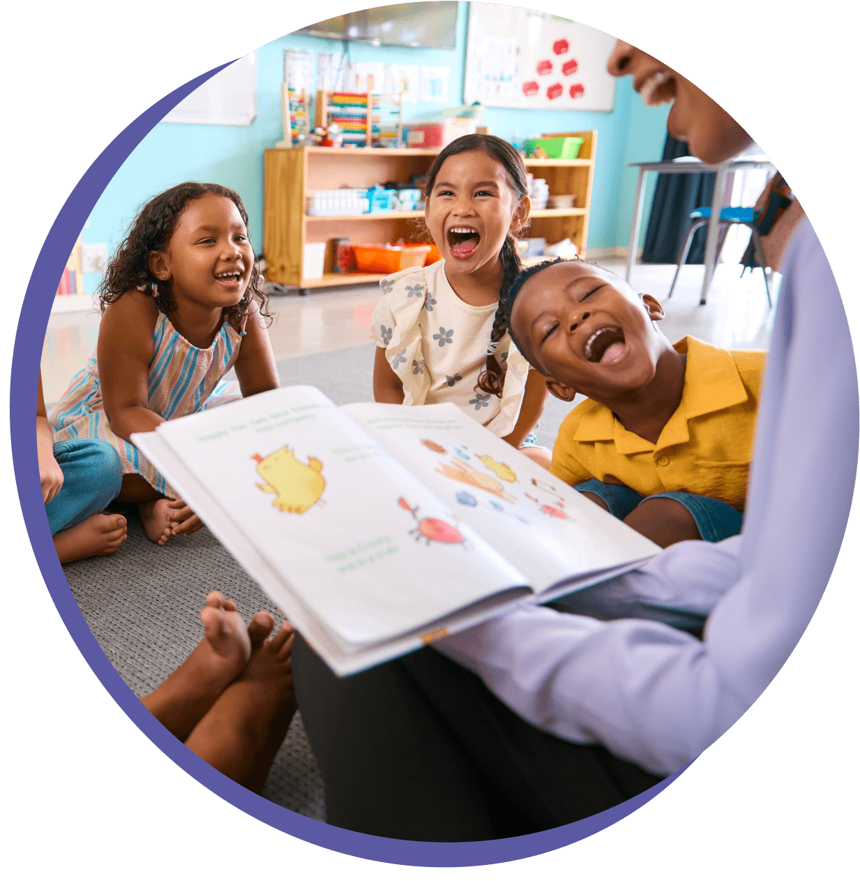 A group of children sit on the floor, laughing, as an adult reads a colorful book to them in a classroom setting.