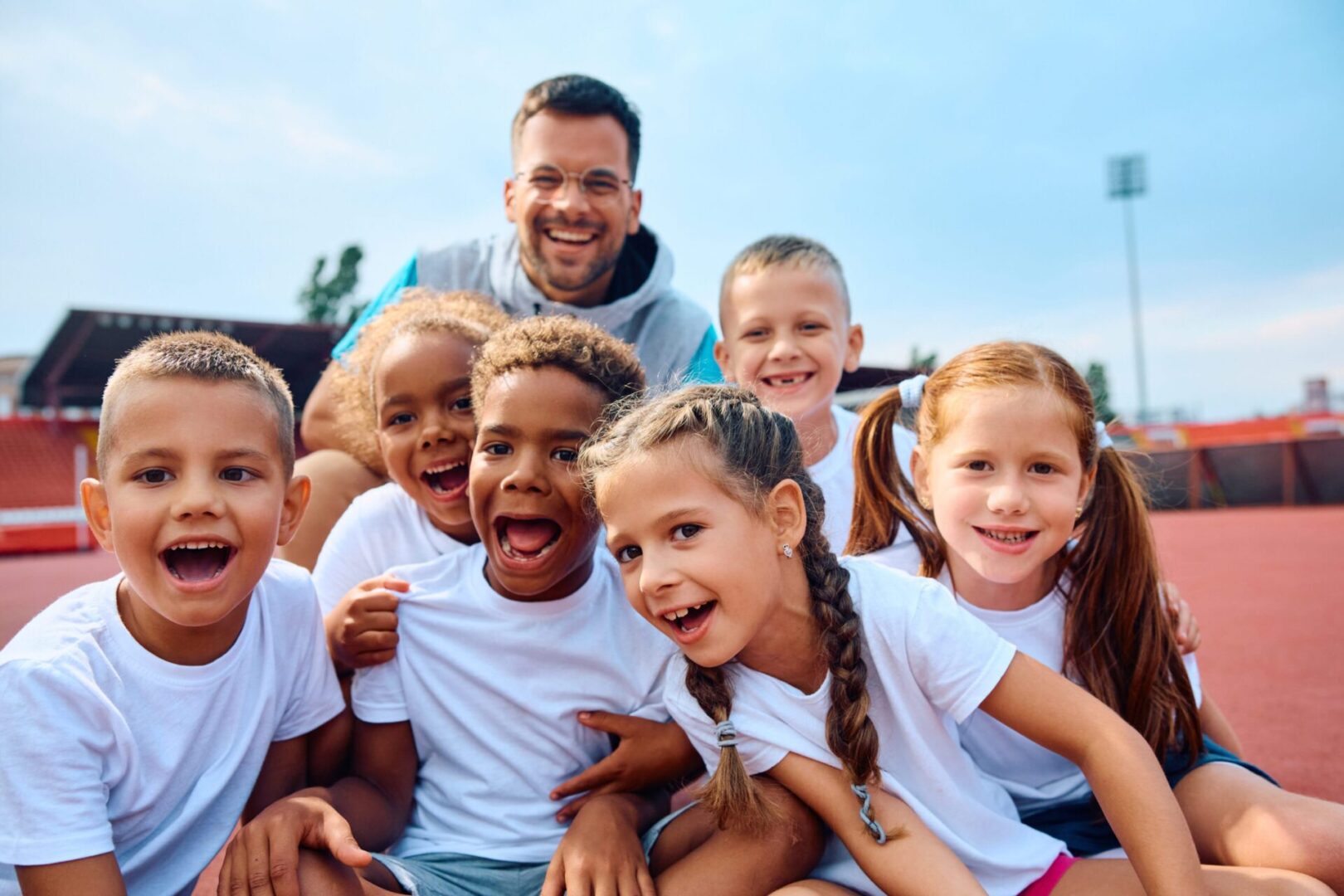 A group of six cheerful children in white shirts and a smiling adult are gathered on a sports field, posing for a photo.