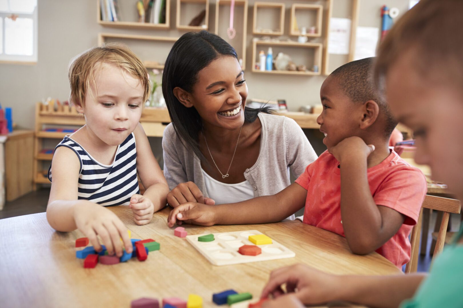 A teacher and three young children interact at a table, engaging in educational activities with colorful blocks and puzzles in a classroom setting.