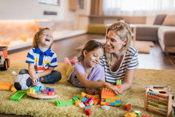 Happy mother and her kids relaxing on carpet at home and playing with toys.